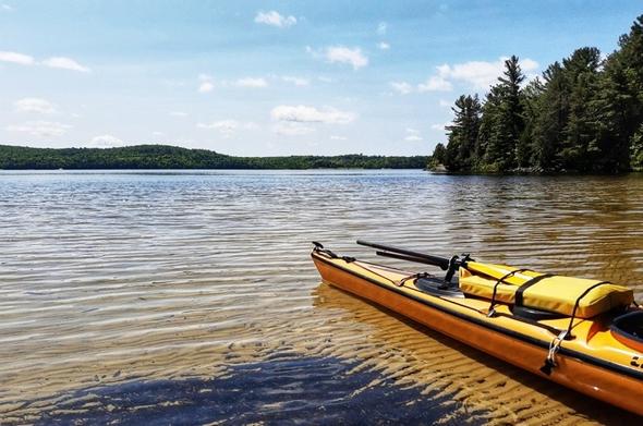 View of a lake with a forested shoreline. A yellow kayak rests on the sand in shallow water.