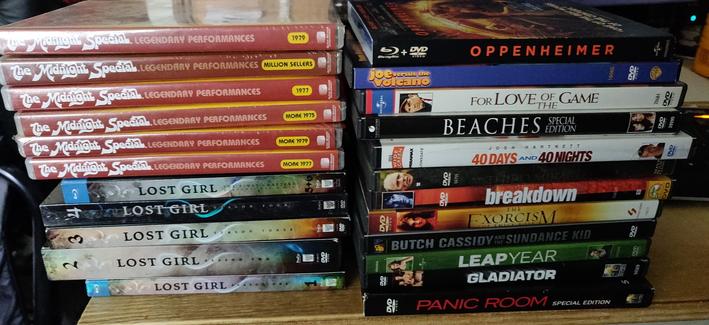 Six Midnight Special collections from the 70s, all six seasons of Lost Girl, 11 DVD movies and Oppenheimer on Blu Ray. Stacked up in two stacks, spines showing, on a table.