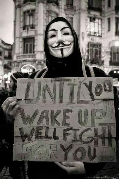 Il cartello nella foto mostra il seguente testo in inglese:

"Until you wake up, we'll fight for you"

La traduzione in italiano è:

"Finché non ti svegli, lotteremo per te"

L'immagine ritrae una persona che indossa la celebre maschera di Guy Fawkes, spesso associata al movimento Anonymous e ad atti di protesta.