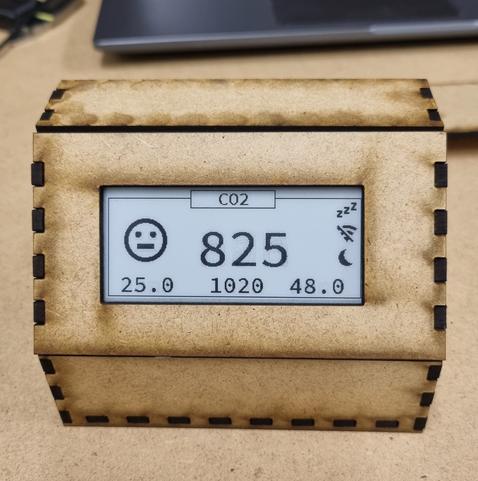 Frontansicht einer Holzbox. Die einzelnen Wände sind mit finger joints ineinander gesteckt. Durch eine Öffnung sieht man ein ePaper Display auf dem oben CO2 steht, groß in der Mitte die Zahl 825, links daneben ein unzufriedenes Emoji. Unten und auf der rechten Seite sind weitere Zahlen und Symbole.
