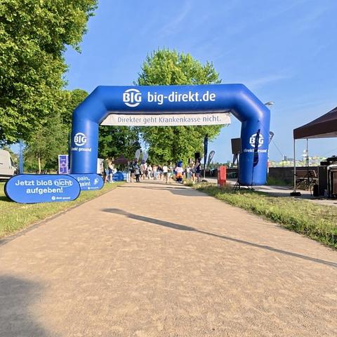 Ein blauer Zielbogen auf dem breiten Fußweg der Rheinwiesen. Einige Menschen und ein paar kleine Zelte (für Getränke?) sind zu sehen. Das war der Start- uns Zielpunkt eines Team-Wander-Events.