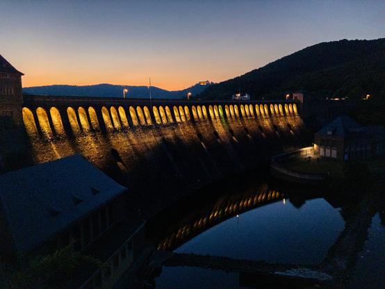 Staumauer am Edersee nach Sonnenuntergang. Der Horizont leuchtet noch orange unter einem Wolkensteins dunkelblauen Himmel. Im Vordergrund dunkel die Staumauer, wobei die schmückenden Torbögen im oberen Bereich farbig - hier gelb - beleuchtet sind. Die Beleuchtung spiegelt sich unterhalb der Staumauer im angestauten Wasser.