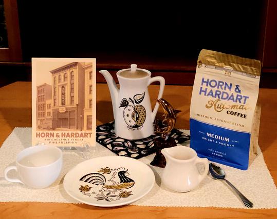 Tea tableau in sepia tones featuring coffee instead of tea, with Horn and Hardart medium blend Automat coffee, a Horn and Hardart postcard, MidCentury Modern coffee pot and plate by Georges Briard with apple and rooster designs, and MidCentury restaurantware from a railroad dining car. There is a shiny bronze glass dolphin on a driftwood pedestal to commemorate the dolphin spigots from which Horn and Hardart coffee flowed.