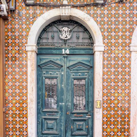 A distressed teal door with two small windows and an arched transom window above, framed by an ornate white archway. The wall surrounding the door is covered in orange, blue, and white Portuguese tiles. The number "14" is visible above the door.