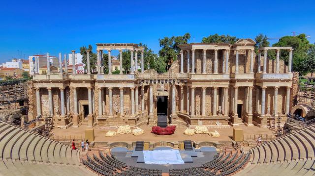 La imatge captura l'antic Teatre Romà de Mérida, Espanya. Les majestuoses ruïnes es troben amb orgull contra un cel blau clar. En primer pla, fileres de seients condueixen a una zona escènica, mentre que les columnes i parets degradades del teatre formen un teló de fons impressionant.