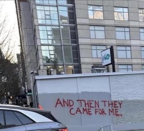 Graffiti reading “AND THEN THEY CAME FOR ME” in red spray paint on a boarded-up wall in an urban area, with a multi-story building and parked cars nearby. The phrase is a reference to the famous anti-fascist poem by Pastor Martin Niemöller, warning against political apathy and the consequences of remaining silent in the face of injustice.