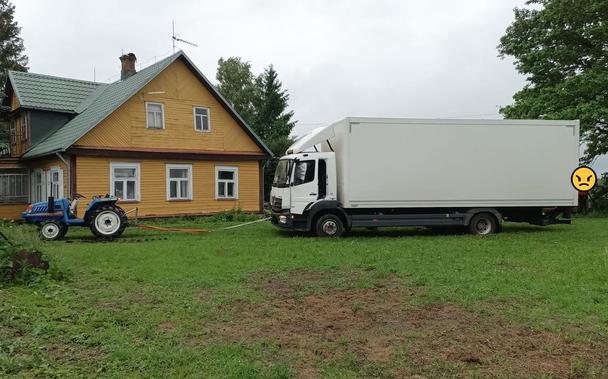 A large white delivery truck, 12 tons I believe, in a grass yard. Its tyres are suspiciously low on the ground. A small blue tractor is attached to the front by some towing strops. A yellow house with green roof is in the background. Behind the truck, a red and black liveried driver is looking angry.