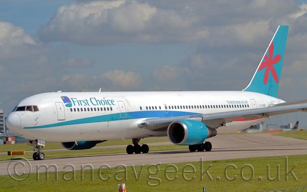 Side view of a twin engined jet airliner taxiing from right to left along a grey runway.
The plane is mostly white, with a multi-tone green/blue stripe running along the body, and green "First Choice" titles on the upper forward fuselage.
There is additional green text "FirstChoice.co.uk" on the upper rear fuselage, and black registration "G-OOBL" just aft of the rear door.
The tail is a green/blue, with a stylised red flower in the middle.
The engine pods under the wing are the same green blue.
Lush green grass lines the runway in the foreground, with pale blue sky and fluffy clouds filling most of the background.