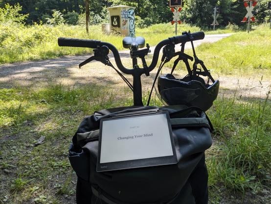 An ebook reader propped up in landscape orientation on the front bag of a brompton bicycle, facing the camera. Centered on the page it reads in large letters: "Part IV Changing Your Mind".

You can see handlebars above the bag with a helmet dangling from it on the right.
The handle bars are curved down in the centre quite a bit (as is typical for Bromptons), allowing a gap for the saddle to be visible through.

Around the bicycle are patches of grass.
Two paths meet behind the bicycle. They're partially in shadows from the forest around this place and partially in bright sun.

In the background you can see the path heading over an unprotected railway crossing with a red light (off).
Next to it is a small beige metal hut that has a black and white Hollow Knight spray on it, aswell as some random graffitti.