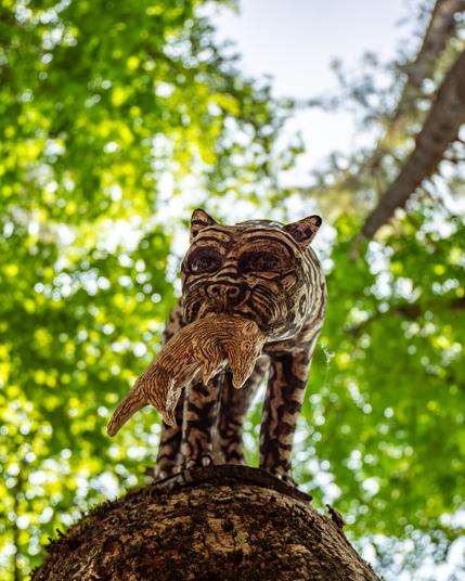 Looking straight up a tree, a striped ceramic cat is attached the tree looking down, The cat has a small rodent (also ceramic) in its mouth. Blur green leaves and other trunks are seen behind the cat