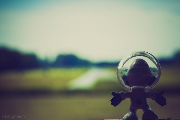 A close-up, shallow depth of field photograph featuring a small, white and blue figurine of an astronaut smurf standing in the foreground. The astronaut figure has a clear, round helmet over its head, and its arms are outstretched. The background is significantly out of focus, showing a blurred outdoor landscape.