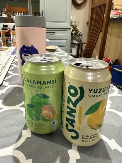 Two cans of Sanzo - left, green can Calamansi with Asian lime pictured on can. Right, Sanzo branding visible. Yellow can. Yuzu label with pic of yuzu fruit. Background, fellow coffee tumbler with blue cat drinking coffee.