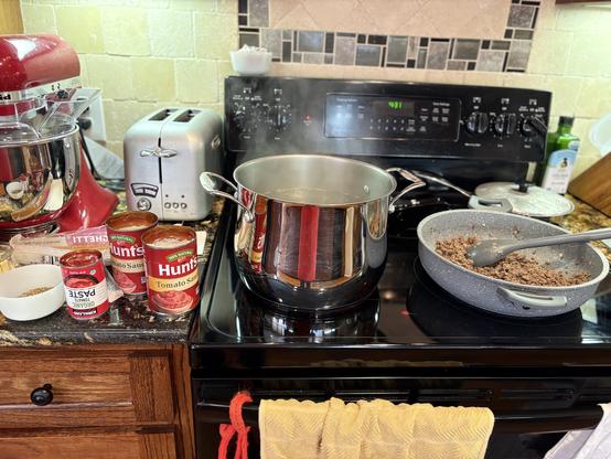 From left to right, on a countertop and a black stovetop: spices in a small white dish; cans of tomato paste and tomato sauce; a pot of boiling water for the pasta, and simmering ground beef with onion and garlic, all ready to be combined for a tasty spaghetti meal.