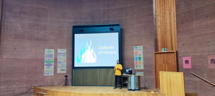 Talk on a wooden stage, presentation in the back reading Cultures of Uncare, in the back is a giant round rammed earth wall