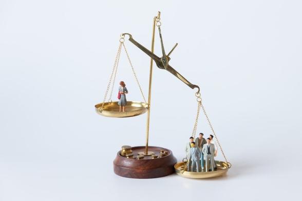 A set of scales with a single woman on the left being outweighed by a group of men on the right. The people are represented using dolls.