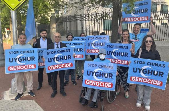 Photo of 10 people holding signs saying "Stop China's Uyghur Genocide". Behind them is the tall metal fence that surrounds the Chinese embassy.