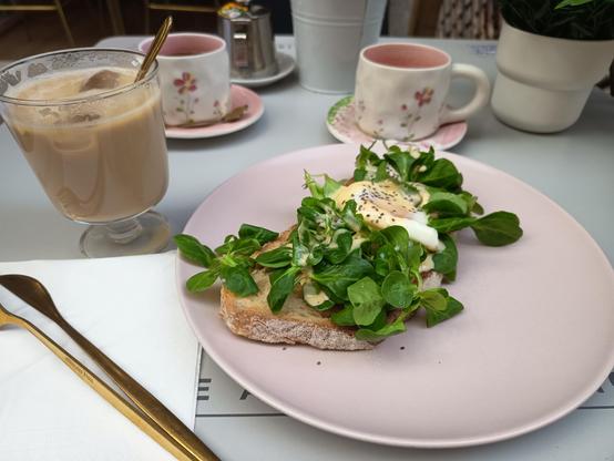 Una fotografía en la que aparecen 2 tazas de fondo con color rosa y dibujos de flores rosas, cada taza tiene su plato rosa. Un vaso con café con leche y hielo y enfrente un plato con una tostada con un huevo escalfado con canónigos y salsa holandesa y semillas de chia por encima.