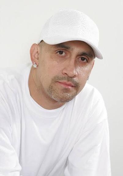 Retrato de Johnny J, un reconocido productor y rapero mexicano, luciendo una camiseta blanca y una gorra blanca, con un fondo neutro.