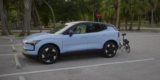 A 2025 Volvo EX30 in cloud blue is parked with a white bicycle behind it. The view looks at the left side of the car. It is in a parking lot with palm trees in the background.