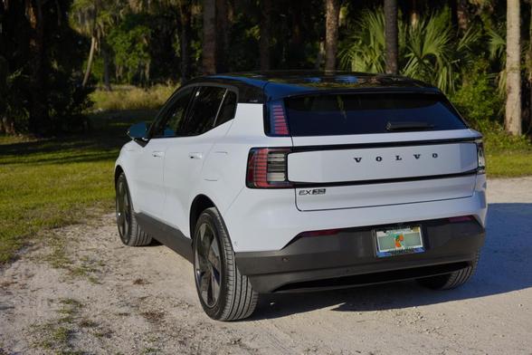 The rear of a 2025 Volvo EX30 in cloud blue, but there is a yellow color cast from the sun getting low on the horizon (not shown). It is parked on sand. There are trees in the background.
