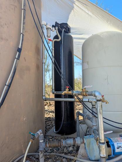 This shows a bit of convoluted galvanized pipe with valves and fittings. On the left is the tan water storage tank, in the middle is a tall black arsenic water filter, and on the right is the well pressure tank. A sunshade is stretched over the whole assembly to protect the plastic parts of the arsenic filter from UV.