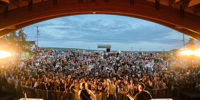 A vibrant photograph of the Yellowknife Folk on the Rocks music festival. A dense crowd of people stands before a stage. The scene takes place outdoors, surrounded by trees and tents under a dusky sky.