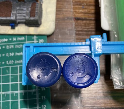 Two tiny bottles of paint displayed bottom-first to show the color difference. The left one is the miscolored bottle, while the right is hopefully correct. Left is significantly darker than right,
Both are held in a vise to ease display. Background is a cutting mat on a table with some other hobby detritus.