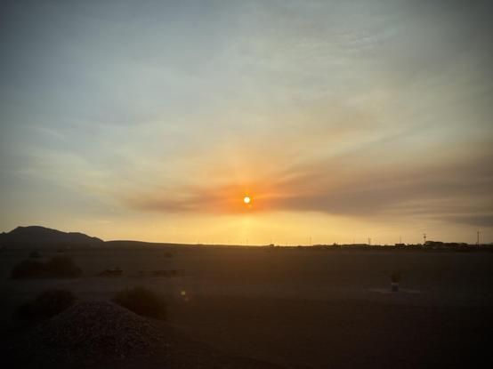 The setting sun shines orange through a wispy cloud of forest fire smoke in a dusky blue sky above the silhouette of the foothills of a desert mountain range at sunset.