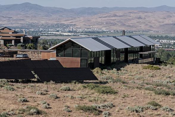 A color landscape photo that shows a very large, long rectangular house with entirely glass walls. The roof is gray colored metal. One of the narrow ends of the house is at center frames that a long side extends out to almost the right side of the frame. In front of the narrow side at center frame are four very large solar arrays, two of which disappear off of the left side of the frame. The house sits in a field of dry grasses and sparse clumps of sagebrush. A city is seen in a wide valley beyond the house. Another large house is seen on the left behind the main house in the photo. On the opposite side of the valley is a mountain range.