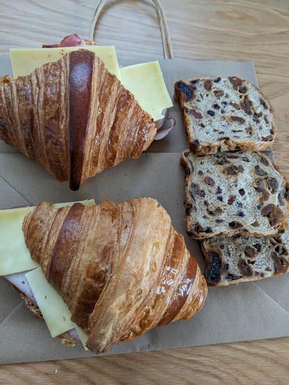 Two large ham and cheese croissant and three slices of fruit loaf sitting on a brown paper bag.