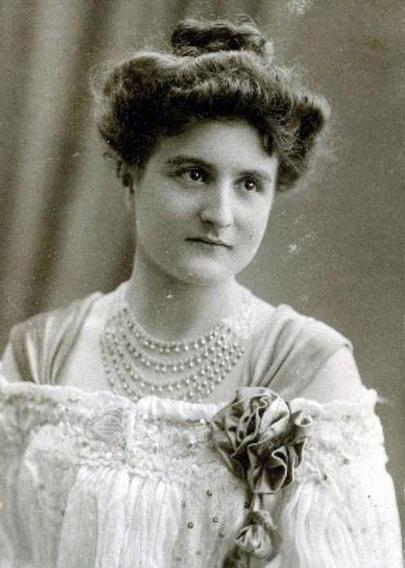 Vintage black-and-white portrait of a woman dressed in an elegant, early 20th-century outfit with detailed embellishments and multiple strands of pearls.