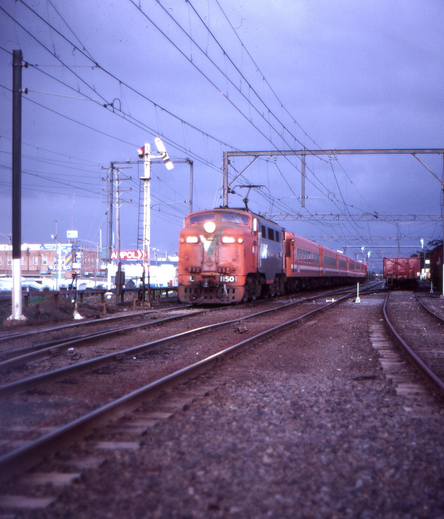 On a dark and stormy late afternoon, in a photo taken from ground level in a railway yard, we see a passenger train in orange livery hauled by an electric locomotive with its headlight shining, approaching us as it passes a somersault semaphore signal in the proceed position.