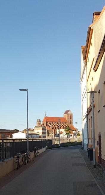 Foto: Wolkenlos blauer Morgenhimmel vor 7:00 Uhr, links Geländer und eine Straßenlaterne an der Unterführung der Poeler Straße unter der Hafenbahn, rechts die letzten Fronten einer Häuserzeile, die vor der Bahnlinie endet. In der Ferne der nördliche Rand des Altstadtkerns, überragt von der mittelalterlichen Backsteinbasilika St. Nikolai und dem Turm der früheren Marienkirche.