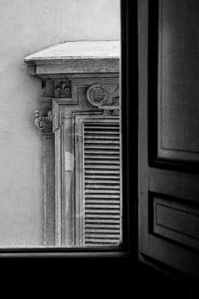 A black and white photo taken through an open window of which you only see the lower right part in the photo. To the right of the window a piece of a wooden shutter, illuminated by the sun. Through the window you see on the other side of the street the upper left corner of a window with a beautifully worked stone edge and part of a wooden shutter with horizontal slats.