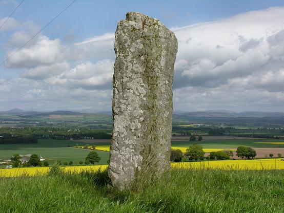 The Keillor Pictish symbol stone. The image shows a grey carved stone in the centre of the frame. It is covered in lichen, but traces of carvings can be made out in it. The stone is set on a hillside that descends behind it past agricultural field to distant hills. The scene is in sunlight.