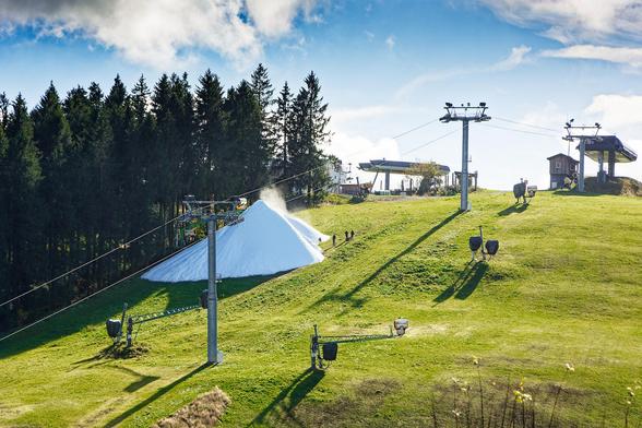 Auf einer grünen sonnenbeschienenen Skipiste wird mit einer Schneelanze ein Haufen Kunstschnee erzeugt.