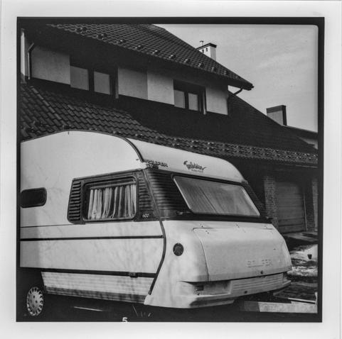 This black and white photograph captures a nostalgic scene featuring an old caravan parked in front of a house with a steep, tiled roof. The caravan has drawn curtains and visible signs of wear, suggesting it has not been moved in some time. The shadow of bare branches falls across its side, adding texture and atmosphere to the image. In the background, the house exhibits a mix of modern and traditional European architectural elements, with visible brickwork, stucco walls, and a chimney. The overall mood is quiet and slightly melancholic, evoking memories of past travels and stillness in the present.