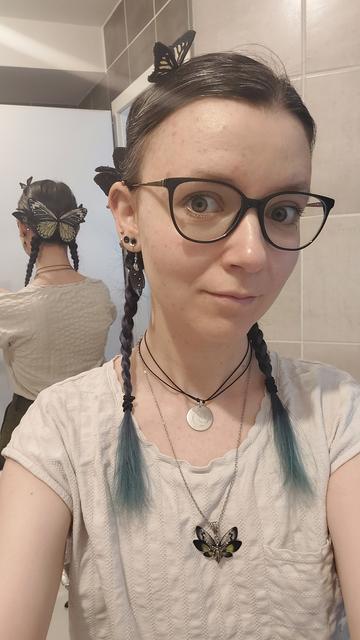 Selfie d'une jeune femme caucasienne, avec des grosses lunettes noires et des nattes brunes aux pointes bleues. 
Elle porte un shirt blanc, un médaillon avec une lune gravée et un pendentif en forme de fée verte. 
Le selfie est pris devant un miroir, ce qui permet de voir son dos. 

A l'arrière de sa tête, elle porte une barrette en forme de grand papillon vert, et plein d'epingles à cheveux sur lesquelles sont cousu d'autres papillons plus petit, et toujours en tissus.