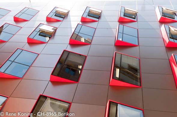 The image shows a modern building facade with a grid of windows. The windows are rectangular and have a distinctive red frame, creating a striking contrast against the gray panels of the building. The windows are arranged in a regular pattern, with some open and others closed, revealing reflections of the sky and neighboring structures. The perspective of the image is from below, looking up at the building, emphasizing the height and design of the windows. The overall color scheme is a combination of gray and red, with the red frames adding a bold visual element to the facade.