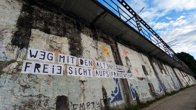 On a wall with graffitis a feminist collage reads: «WEG MIT DEN ALTEN [WEISSEN MÄNNERN] - FREIE SICHT AUFS FRAUENMEHR» translates to: «GET RID OF OLD [WHITE MEN] - FOR A CLEAR VIEW ON A WOMEN'S MAJORITY». On the right top corner a view of a blue cloudy sky.