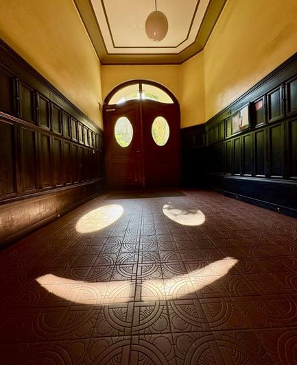 A brightly lit hallway featuring a decorative patterned floor and wooden paneling. The focus is on two light spots cast on the floor, resembling a smile. Large double doors with oval windows are visible at the end of the corridor, illuminated by natural light.