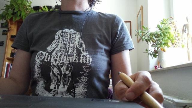 A person is sitting at a desk. The torso can be seen wearing a gray T-shirt by the band Obelyskkh, with a white print of a headless figure in medieval Far Eastern garb, with long white threads flowing from their hands and lying at their feet. The band name is in the middle of the figure.