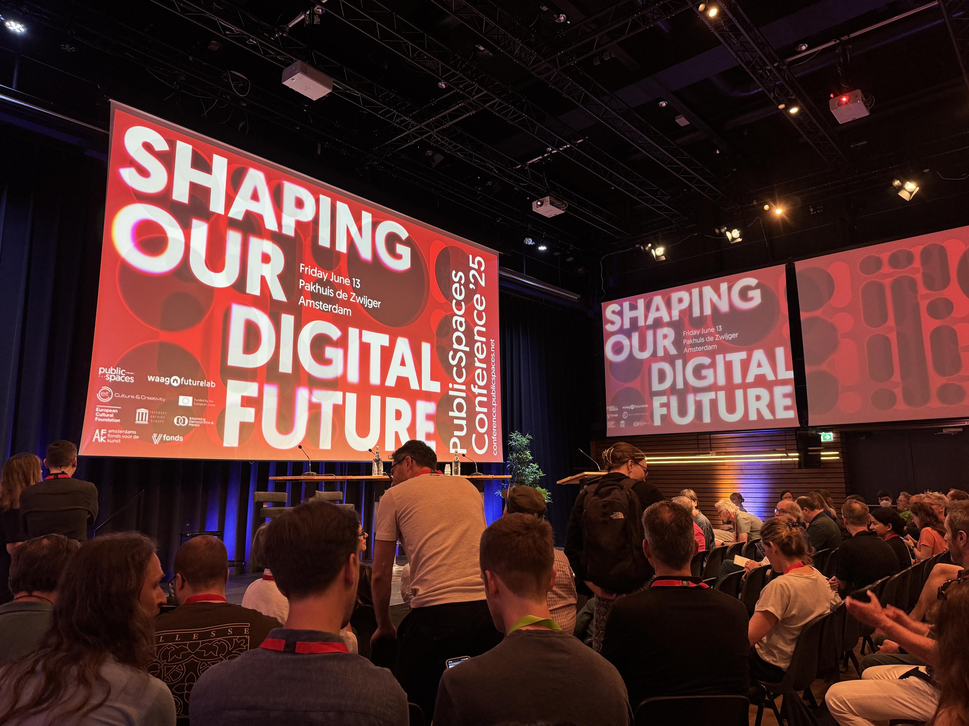 Tech conference “Shaping Our Digital Future” at Pakhuis de Zwijger, Amsterdam. Packed auditorium with large red screens displaying the event branding.​​​​​​​​​​​​​​​​