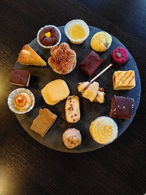 Auf einer großen, runden Schieferplatte thronen  diverse süße Köstlichkeiten im Kleinstformat: Plätzchen, Törtchen, Brownies, Cupcakes, manche mit Schokolade, manche mit Früchten, andere mit Crèmes oder Nüssen. Manche stecken in kleinen Papierförmchen. Es gibt runde und eckige Leckereien.