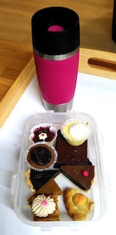 Auf einem Tablett stehen ein von einer Gästin mitgebrachten pinker Thermobecher mit schwarzem Deckel und davor eine rechteckige Kunststoffschale mit vielen kleinen Kuchenstücken. Der Schwerpunkt liegt hier auf schokoladigen Genüssen: Shortbread, Brownies, Törtchen, mal cremig, mal fest, mit Nüssen oder pur - auf jeden Fall immer lecker!