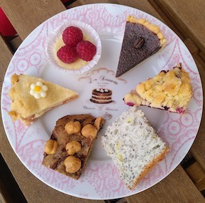 Ein Kuchenteller mit filigran verziertem, rosafarbenen Rand, auf dem sechs verschiedene Leckereien im Kleinstformat (im Uhrzeigersinn): Vollmilchschokoladen-Espresso-Törtchen, Beeren-Streusel-Törtchen, Angel Food Cake mit Zitrone und Mohn, Schoko-Haselnuss-Keks, gedeckter Pfirsichkuchen und ein Cheesecake mit Himbeeren aus eigenem Anbau.
