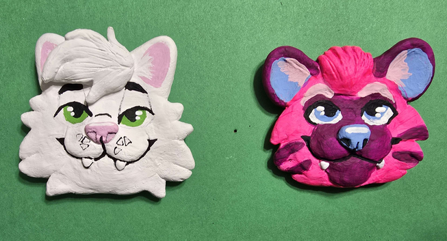 two clay headshot pins, one of a white cat with green eyes, the other of a neon pink hyena with blue eyes and nose