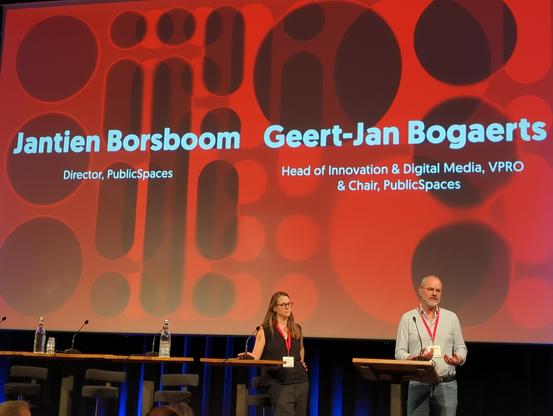 Big screen on stage with redish background showing the texts:
Jantien Borsboom Director, PublicSpaces Geert-Jan Bogaerts,  Head of Innovation & Digital Media, VPRO & Chair, PublicSpaces.

Jantien and Geert-Jan are standing in from of the screen