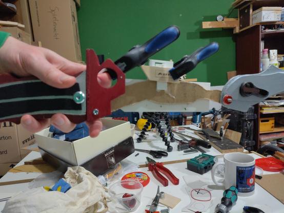 Hand holding a handle with a red part on it, which is connected to a brown and a white part, which is connected to a silver part. The parts form a horizontal line and are held in the air. Clamps are attached to the white part, there's hot glue all over it. Clearly a prototype. Background is a workbench full of tools, a coffee mug and a box with cosplay stuff.