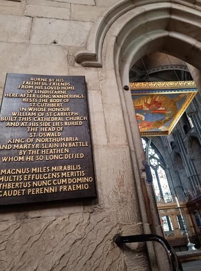 The sign reads:
Borne by his faithful friends from his loved home of Lindisfarne here after long wanderings rests the body of St Cuthbert in whose honour William of St Carileph built this cathedral church and at his side lies buried the head of St Oswald king of Northumbria and martyr slain in battle by the heathen whom he so long defied

(There follows an inscription in Latin)
